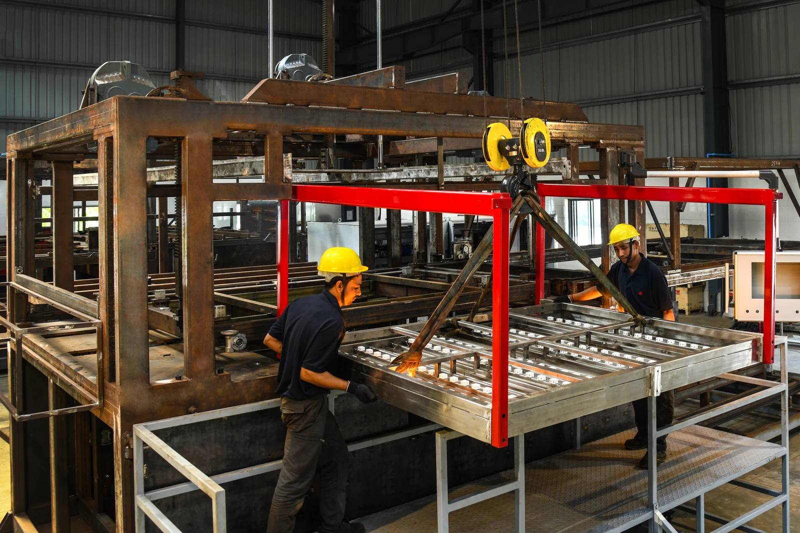 Workers using a chain hoist to position a large assembly inside an unfinished machine frame.